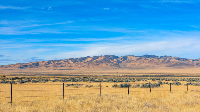Barbed wire fence crossing dry grassland in Nevada desert landscape with mountains and blue sky