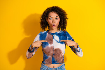 Surprised young woman points at herself against a bold yellow background wearing stylish tie-dye top
