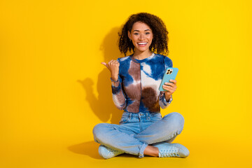 Happy young woman with curly hair holding a smartphone while seated on a yellow background wearing casual modern style