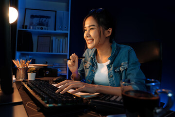 Smiling young beautiful asian creative officer looking on pc in sales target online, earning job's...