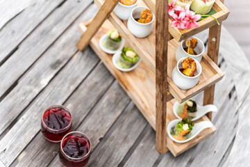 Minimal three-tier wooden stand displaying assorted appetizers and beverages by a poolside. Relaxing outdoor setting with natural light and greenery in the background.