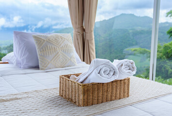 Rolled white towels in a woven basket placed on a cozy bed with mountain view through floor-to-ceiling glass. Serene luxury retreat in nature. Rainy season.