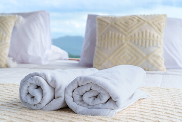 Rolled white towels on a cozy bed with mountain view through floor-to-ceiling glass in natural sun light. Serene luxury retreat in nature. Rainy season.