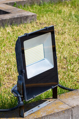 A black flood light is positioned on a brick wall among the grass