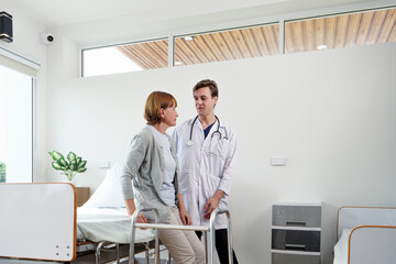Caucasian doctor supporting elderly female patient using walker in bright hospital room, guiding rehabilitation steps near bed, symbolizing professional care and patient recovery.