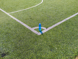 A blue sports water bottle stands in the corner of an artificial turf soccer field. The clear white...
