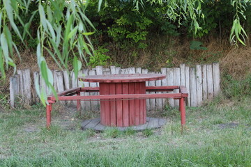 A red wooden picnic table surrounded by greenery and wooden fencing.
