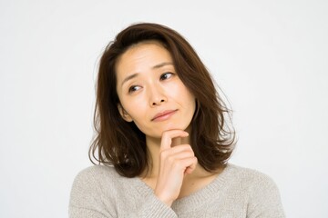 Thoughtful Asian woman with shoulder-length brown hair wea casual gray sweater ponde with hand on chin against plain white background