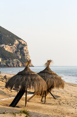 Two straw beach umbrellas on a sandy shore with rocky cliff and calm sea in the background at sunset. Peaceful coastal scenery and natural beach setting. Outdoor vacation and summer atmosphere.
