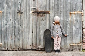 Cute little girl in winter wool clothes with skateboard while standing against grunge wooden background - Concept of activity and playful childhood - Outdoor games in courtyard - Copy promo space