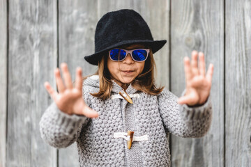 playful young girl in sunglasses and black hat gestures with hands in front of a rustic wooden wall. Her expression mixes fun and confidence. Generation alpha childhood and lifestyle concept.