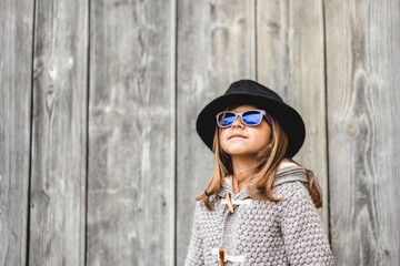 Confident child girl wearing a black hat and blue sunglasses poses in front of a rustic wooden wall. Her style and expression convey individuality and self-confidence. Generation alpha concept.