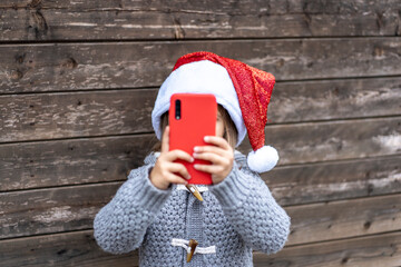 Little girl wearing Santa Claus hat having fun with a red smartphone outdoors during Christmas time - Little girl standing against a wooden  background - E-commerce and tech concept for Xmas gifts