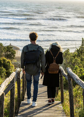 Young couple walking hand in hand on a wooden path toward the beach with ocean waves in the distance. Back view in natural light. Romantic moment and travel lifestyle concept.