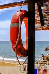 Red lifebuoy hanging on a wooden beach hut with the sea and sandy shore in the background. Bright summer day with strong sunlight and beach atmosphere. Safety and vacation concept.