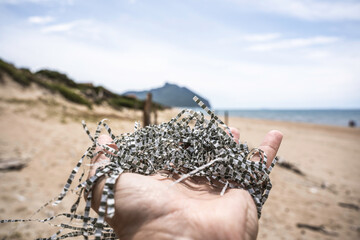 Hand holding shredded microplastic strips on a wild sandy beach with a mountain in the background. Symbol of environmental pollution and human impact on natural ecosystems.