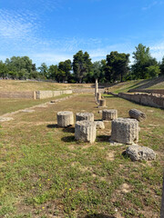 Ruins of an ancient site with scattered stone columns in a grassy arena beneath a bright blue sky