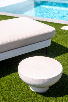 Modern white sun lounger and round table on artificial grass beside turquoise pool under sunlight, creating minimalist luxury outdoor living and contemporary Mediterranean architecture elegance.