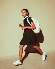 Young girl in school uniform running with backpack on beige background