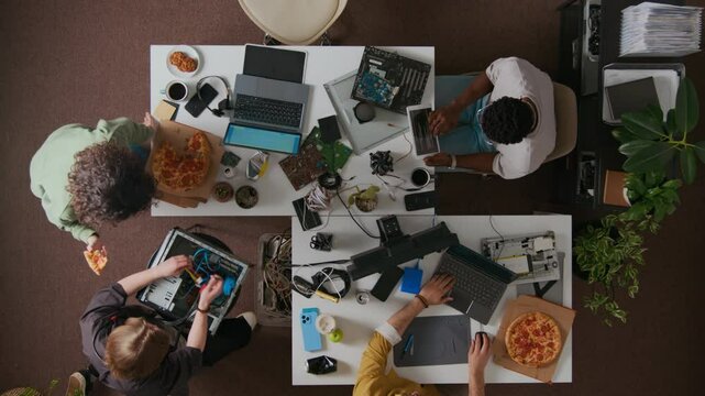 Directly above shot of girl with pizza slice checking cooling fan leaning over seated man connecting PC case cables, two male coworkers using tablet and laptop at desks