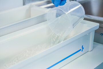 Close-up of a gloved hand pouring clear liquid from a transparent measuring cup into a white tray,...
