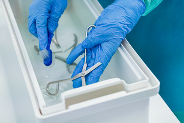 Gloved hands diligently clean various stainless steel instruments with a small brush in a white...