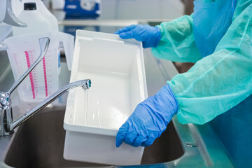 Worker in protective attire and blue gloves thoroughly rinses a white laboratory container in a...