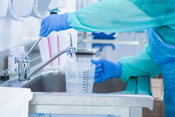 Hands in blue protective gloves filling a clear measuring cup with water from a faucet in a sterile...
