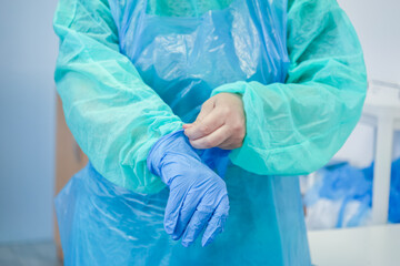 A medical professional's hands in a green gown carefully pull on a blue latex glove, emphasizing...