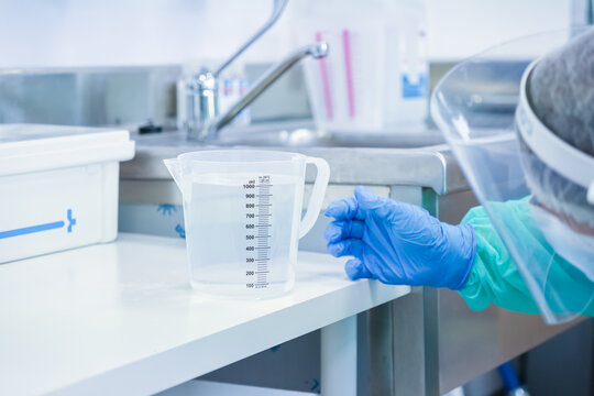Professional in PPE and blue gloves next to a measuring jug of liquid, highlighting meticulous care and precise measurements within a lab environment