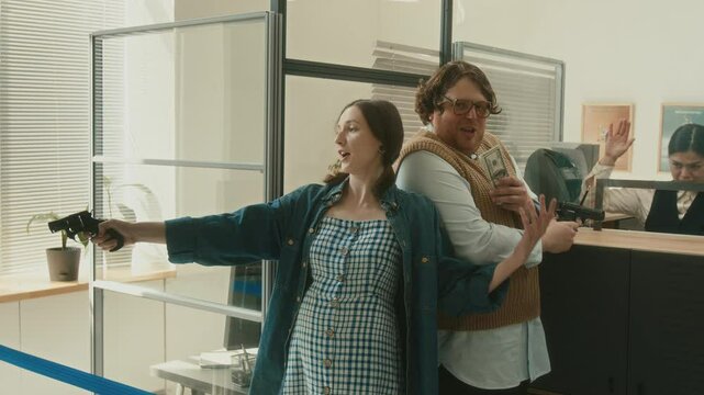 Medium shot of unprofessional male criminal robbing bank with pregnant female partner pointing gun at hostages while excited man holding wad of cash at tellers counter