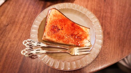 close up of a slice of baked basque cheese cake looks like creme brulee custard cake with caramelized top with a glossy, crackled surface on an elegant plate with two ornate silver forks
