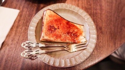 close up of a slice of baked basque cheese cake looks like creme brulee custard cake with caramelized top with a glossy, crackled surface on an elegant plate with two ornate silver forks