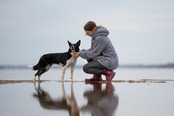 A woman with a hooded jacket is crouched on a shore, petting her black and white Border Collie. The...