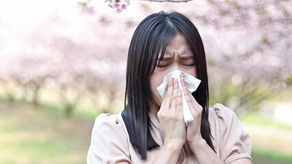 Young asian woman sneezing into tissue under blooming cherry trees, spring allergy or hay fever concept. Seasonal health, nature, and allergy awareness outdoors. Copy space for text