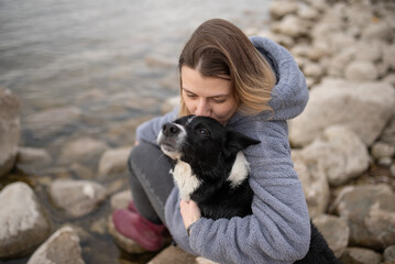 A woman in a hooded sweatshirt lovingly embraces her black and white Border Collie on a rocky beach...