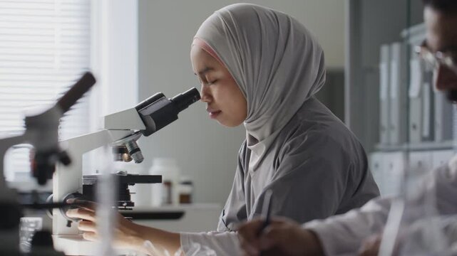 Female scientist in hijab observing slide through microscope while sitting beside male colleague taking notes at desk during research in laboratory