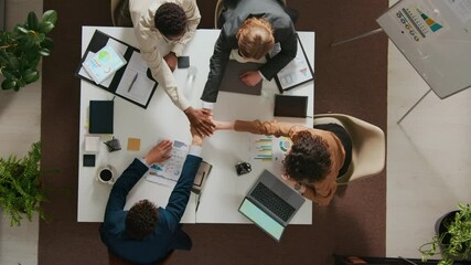 High angle shot of positive team of four diverse coworkers stacking hands and celebrating success while sitting at white table in office setting - Powered by Adobe