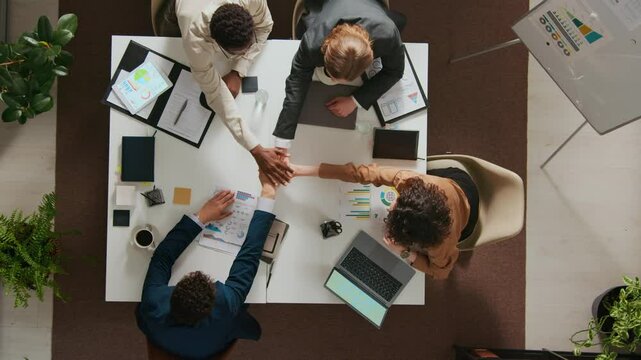 High angle shot of positive team of four diverse coworkers stacking hands and celebrating success while sitting at white table in office setting