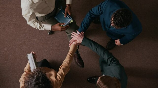 View from above of four multiethnic young people in formalwear stacking hands supporting team spirit in office with brown carpet on floor