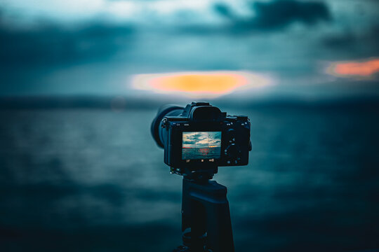A close-up shot of a digital camera on a tripod, capturing a serene lake sunset with dramatic blue and orange tones in Switzerland. - Powered by Adobe