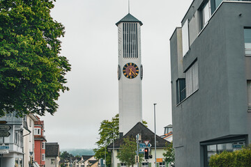 A tall modern clock tower with a minimalist design stands between residential buildings in a small...