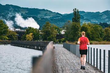 Man in red shirt jogging on a scenic wooden bridge over the lake, with lush green trees and hills...