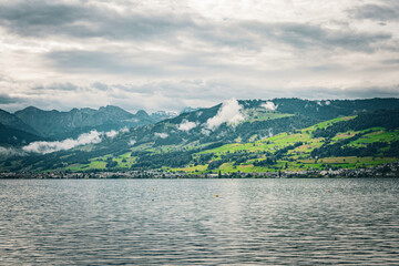 Panoramic view of Swiss Alpine foothills with vibrant green pastures, small town on the lakeshore,...