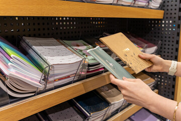 A person selects a notebook with NOTE printed on its cover from a diverse stationery shelf in a...