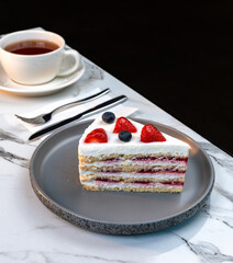 Layered Vanilla Berry Cake Slice with Cream and Fresh Fruits Served with Hot Tea in White Cup