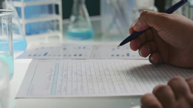 Hands of scientist filling out laboratory research documentation at desk surrounded by chemical glassware. Close-up shot