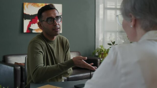 Medium full shot of adult Indian man talking to psychologist exploring emotions through dialogue with specialist during private practice at counselling office