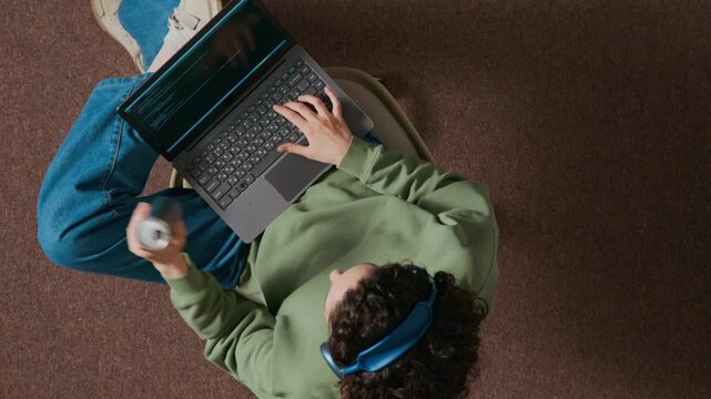 High angle view of curly-haired female programmer wearing headphones sitting on chair with laptop on her lap, writing code and drinking beverage holding can in office