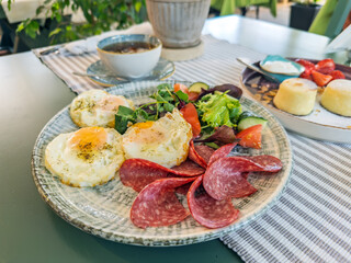 A delicious and hearty breakfast plate with fried eggs, salami, and fresh vegetable salad. A complete morning meal served in a cafe